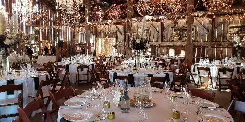 A rustic event venue interior featuring multiple round tables set with white linens, glassware, and centerpieces, surrounded by wooden chairs and string lights.