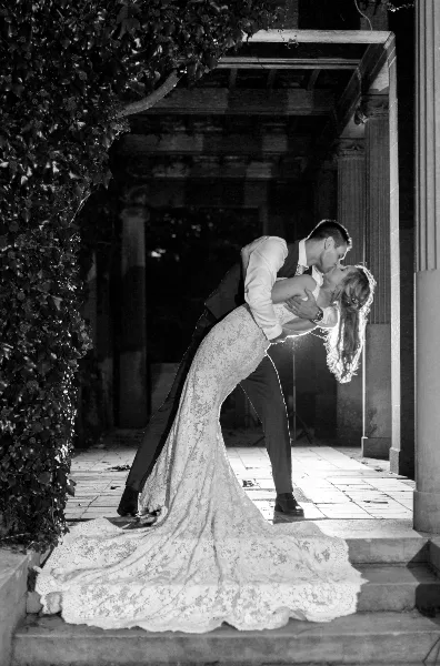 Black-and-white photo of a groom dipping his bride in a lace gown as they kiss on stone steps beside an ivy-covered wall.
