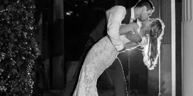 Black-and-white photo of a groom dipping his bride in a lace gown as they kiss on stone steps beside an ivy-covered wall.