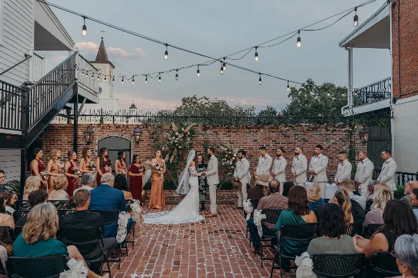 Outdoor wedding ceremony in a brick courtyard with string lights and a floral arch at a historic venue.