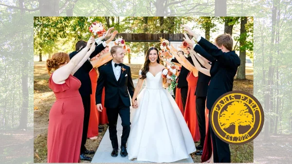 A bride and groom walking down a concrete path lined by wedding party members in a wooded outdoor venue.
