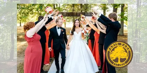 A bride and groom walking down a concrete path lined by wedding party members in a wooded outdoor venue.
