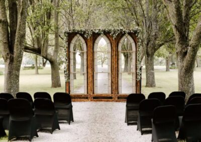 Outdoor wedding ceremony setup featuring a wooden arched backdrop with floral draping and rows of black chairs.