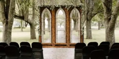 Outdoor wedding ceremony setup featuring a wooden arched backdrop with floral draping and rows of black chairs.