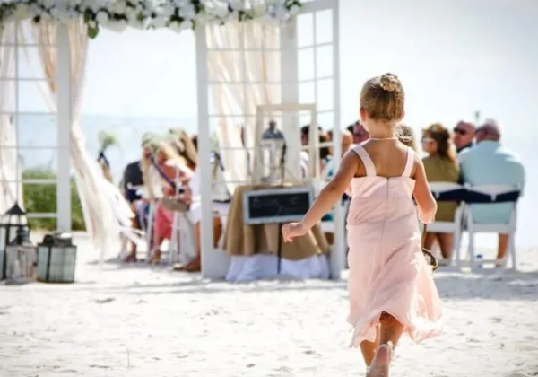 A flower girl in a pink dress walks toward a wedding ceremony arch on a beach with guests seated in the background.