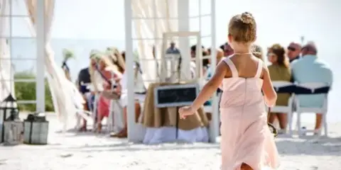 A flower girl in a pink dress walks toward a wedding ceremony arch on a beach with guests seated in the background.