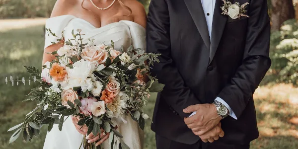 A bride and groom standing outdoors, with the bride holding a large bouquet of white, peach, and pink flowers and the groom wearing a matching boutonniere.
