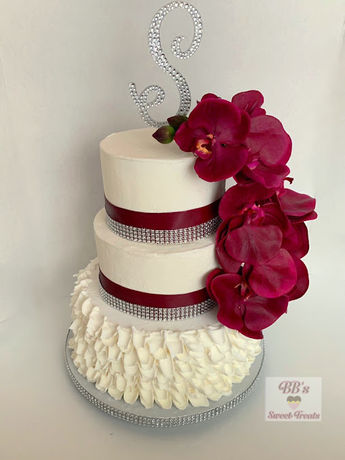 A three-tier white wedding cake decorated with burgundy ribbons, silver beaded trim, and a cascade of red orchids.