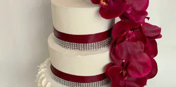 A three-tier white wedding cake decorated with burgundy ribbons, silver beaded trim, and a cascade of red orchids.
