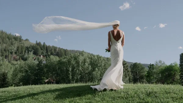 A bride in a white wedding dress stands on a grassy hill with her veil blowing in the wind, set against a backdrop of pine trees and a blue sky.