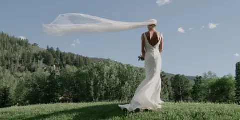 A bride in a white wedding dress stands on a grassy hill with her veil blowing in the wind, set against a backdrop of pine trees and a blue sky.