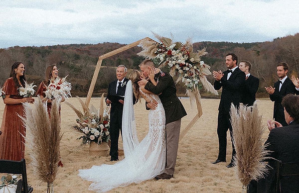 A bride and groom kissing during an outdoor wedding ceremony in a field with a wooden arch and floral arrangements.