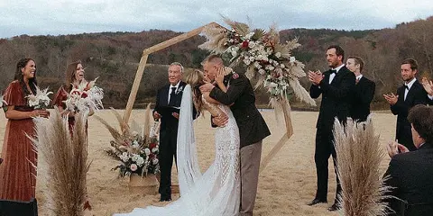 A bride and groom kissing during an outdoor wedding ceremony in a field with a wooden arch and floral arrangements.