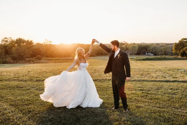 A bride and groom dancing in a grassy field during a sunset.