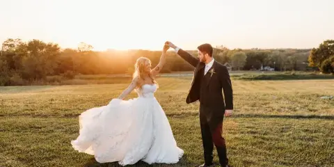 A bride and groom dancing in a grassy field during a sunset.