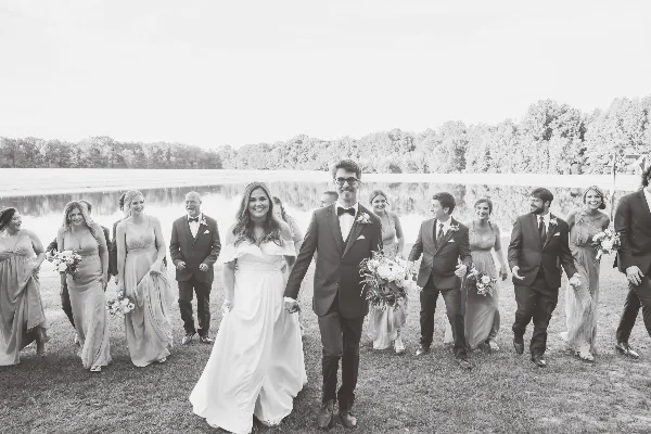 A black and white photograph of a bride and groom walking with their wedding party on a grassy area next to a pond.