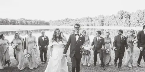 A black and white photograph of a bride and groom walking with their wedding party on a grassy area next to a pond.