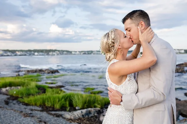 A bride and groom sharing a kiss on a rocky shoreline with the ocean in the background.