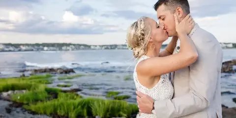 A bride and groom sharing a kiss on a rocky shoreline with the ocean in the background.