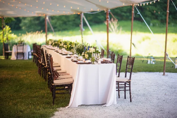 A long wedding reception table set under a tent with white linens, chairs, and floral centerpieces.