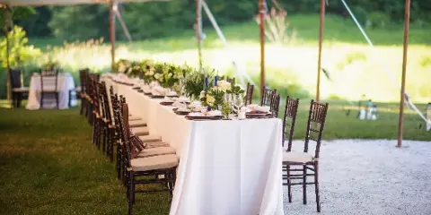 A long wedding reception table set under a tent with white linens, chairs, and floral centerpieces.