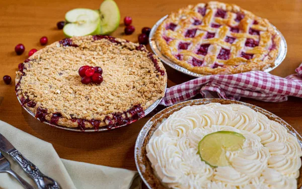 Three freshly baked pies on a wooden table, including a crumb-topped berry pie, a lattice-topped pie, and a cream pie with a lime slice, garnished with apples and cranberries.