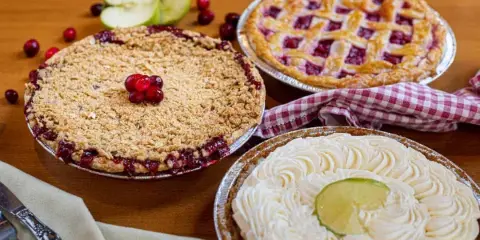 Three freshly baked pies on a wooden table, including a crumb-topped berry pie, a lattice-topped pie, and a cream pie with a lime slice, garnished with apples and cranberries.