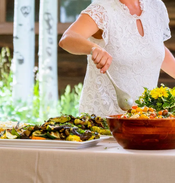 A person in a white lace top serving roasted vegetables and a salad from a wooden bowl at an event table.
