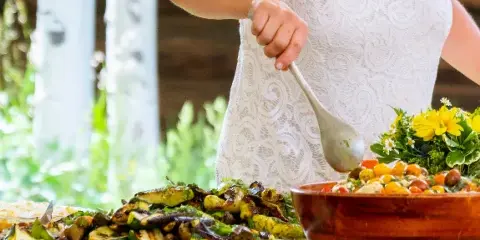 A person in a white lace top serving roasted vegetables and a salad from a wooden bowl at an event table.