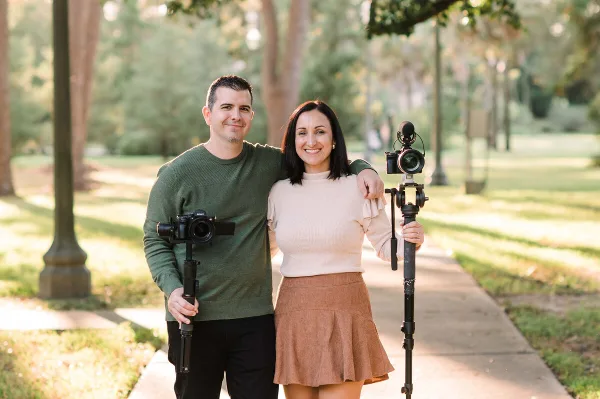 A man and a woman standing together outdoors, holding professional video cameras and stabilizers.