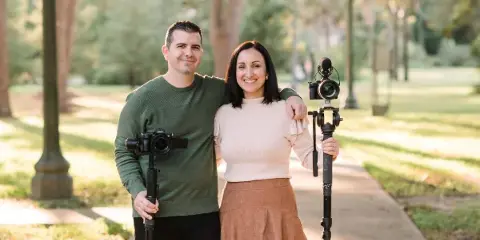 A man and a woman standing together outdoors, holding professional video cameras and stabilizers.
