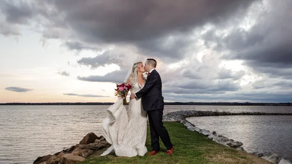 A bride and groom sharing a kiss on a grassy point overlooking a body of water under a dramatic, cloudy sky.