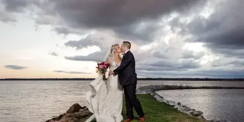 A bride and groom sharing a kiss on a grassy point overlooking a body of water under a dramatic, cloudy sky.
