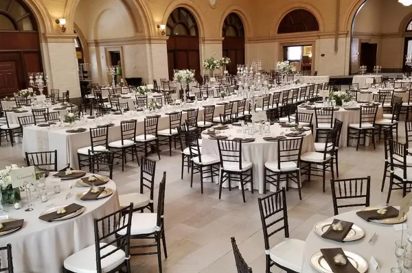 A wedding reception hall set up with round tables, white tablecloths, and crossback chairs.