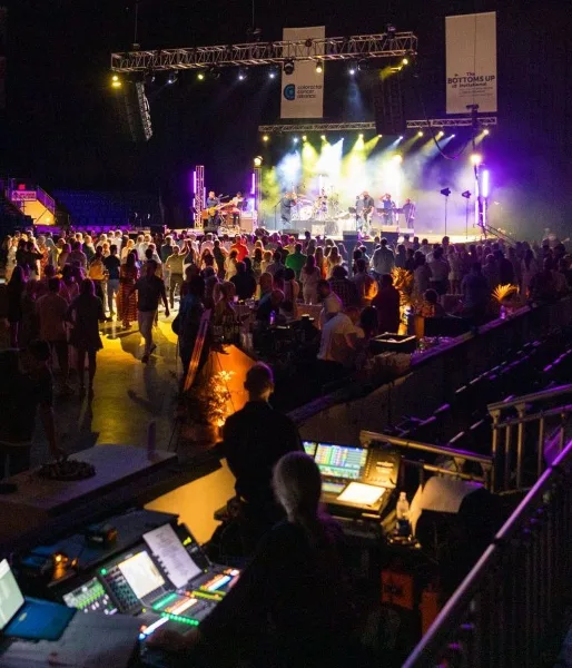 Live event production scene featuring a stage with performers, stage lighting, and a soundboard in the foreground.