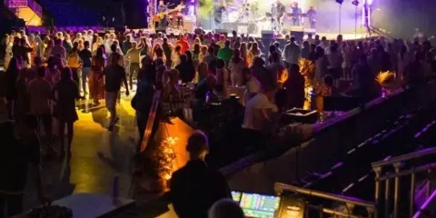 Live event production scene featuring a stage with performers, stage lighting, and a soundboard in the foreground.