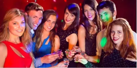 A group of smiling young adults holding shot glasses in a dimly lit room with colorful party lighting.