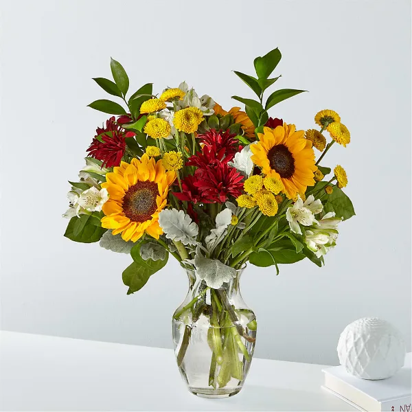 A mixed bouquet of sunflowers, red dahlias, yellow chrysanthemums, and white alstroemeria in a clear glass vase.