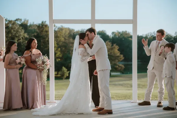 A bride and groom share a kiss during an outdoor wedding ceremony under a white structure, surrounded by bridesmaids and groomsmen.