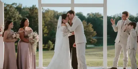 A bride and groom share a kiss during an outdoor wedding ceremony under a white structure, surrounded by bridesmaids and groomsmen.