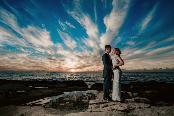 A bride and groom standing on a rocky shore facing each other against a dramatic sunset sky and ocean horizon.