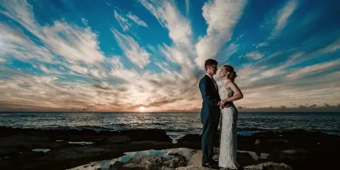 A bride and groom standing on a rocky shore facing each other against a dramatic sunset sky and ocean horizon.