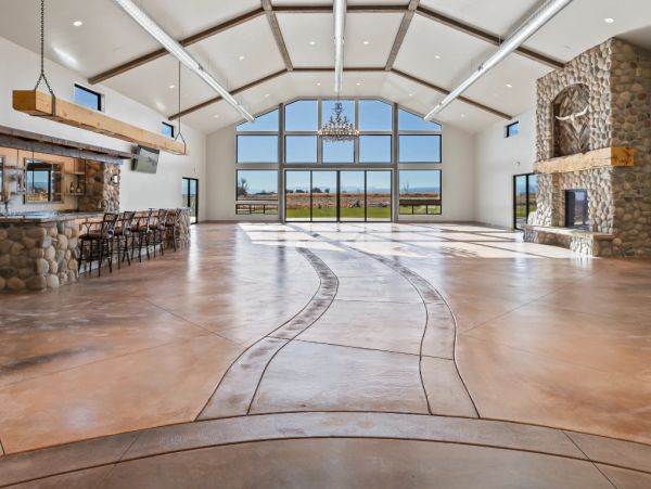 Interior view of a spacious wedding venue with a vaulted ceiling, stone fireplace, bar area, and large windows overlooking a field.