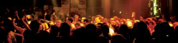 A crowd of people at a party or event with hands raised, illuminated by warm stage lighting.