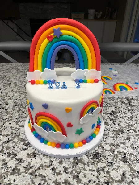 A white custom cake decorated with a large rainbow arch, clouds, and colorful sprinkles.