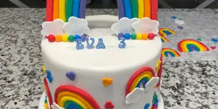 A white custom cake decorated with a large rainbow arch, clouds, and colorful sprinkles.
