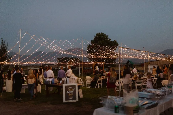 An outdoor evening event with string lights, tents, and guests seated at tables.