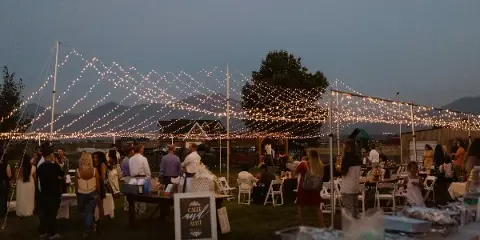 An outdoor evening event with string lights, tents, and guests seated at tables.