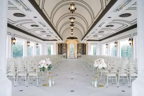 Interior view of a wedding venue featuring white chairs, floral centerpieces, and an ornate ceiling.