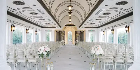 Interior view of a wedding venue featuring white chairs, floral centerpieces, and an ornate ceiling.
